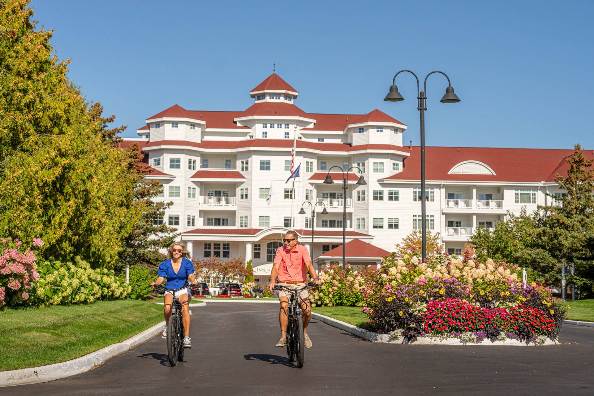 Couple in sunglasses ride bikes down front drive of Inn at Bay Harbor alongside colorful gardens on summer day