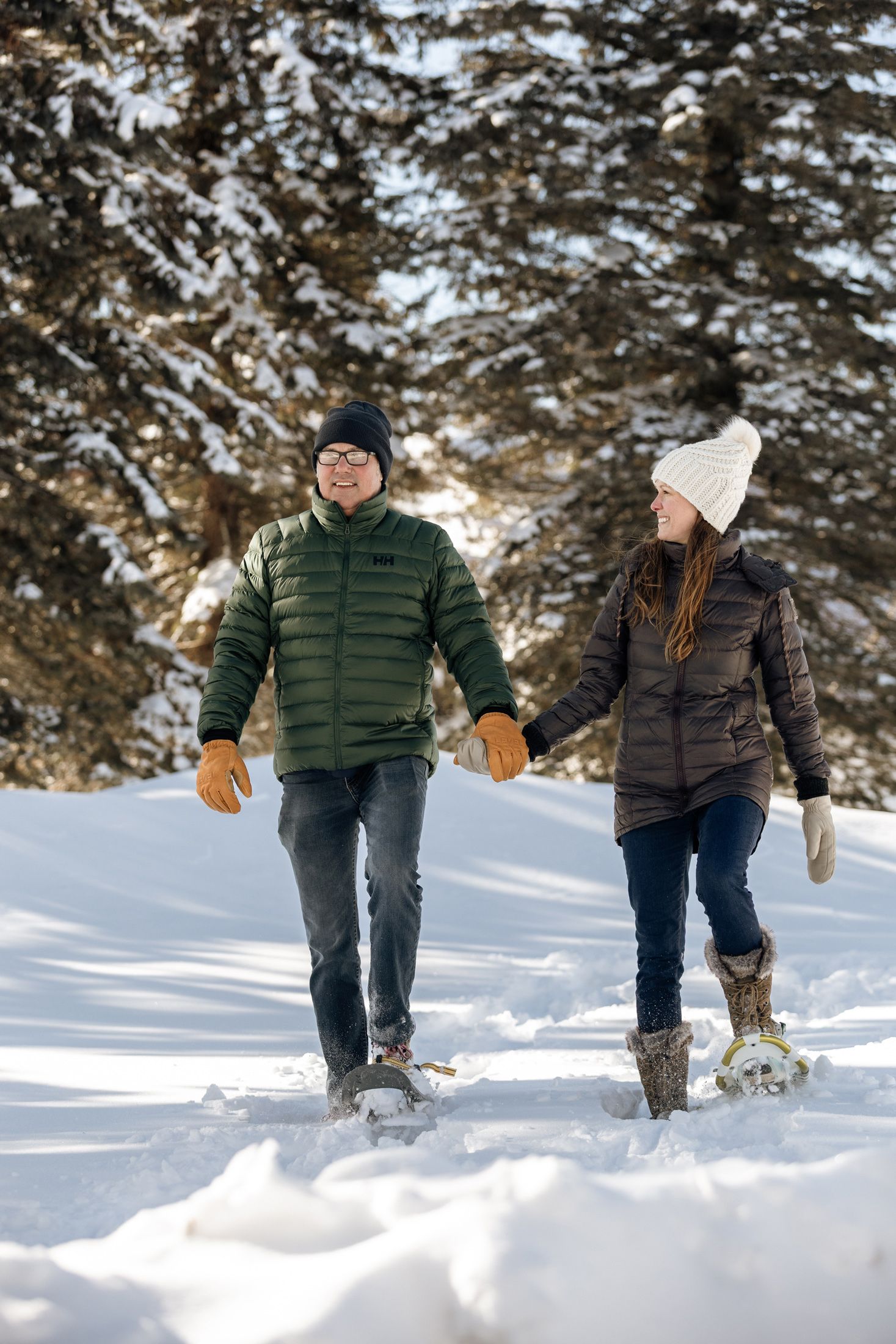 Winter-clad couple snowshoes down snowy trail with pine trees