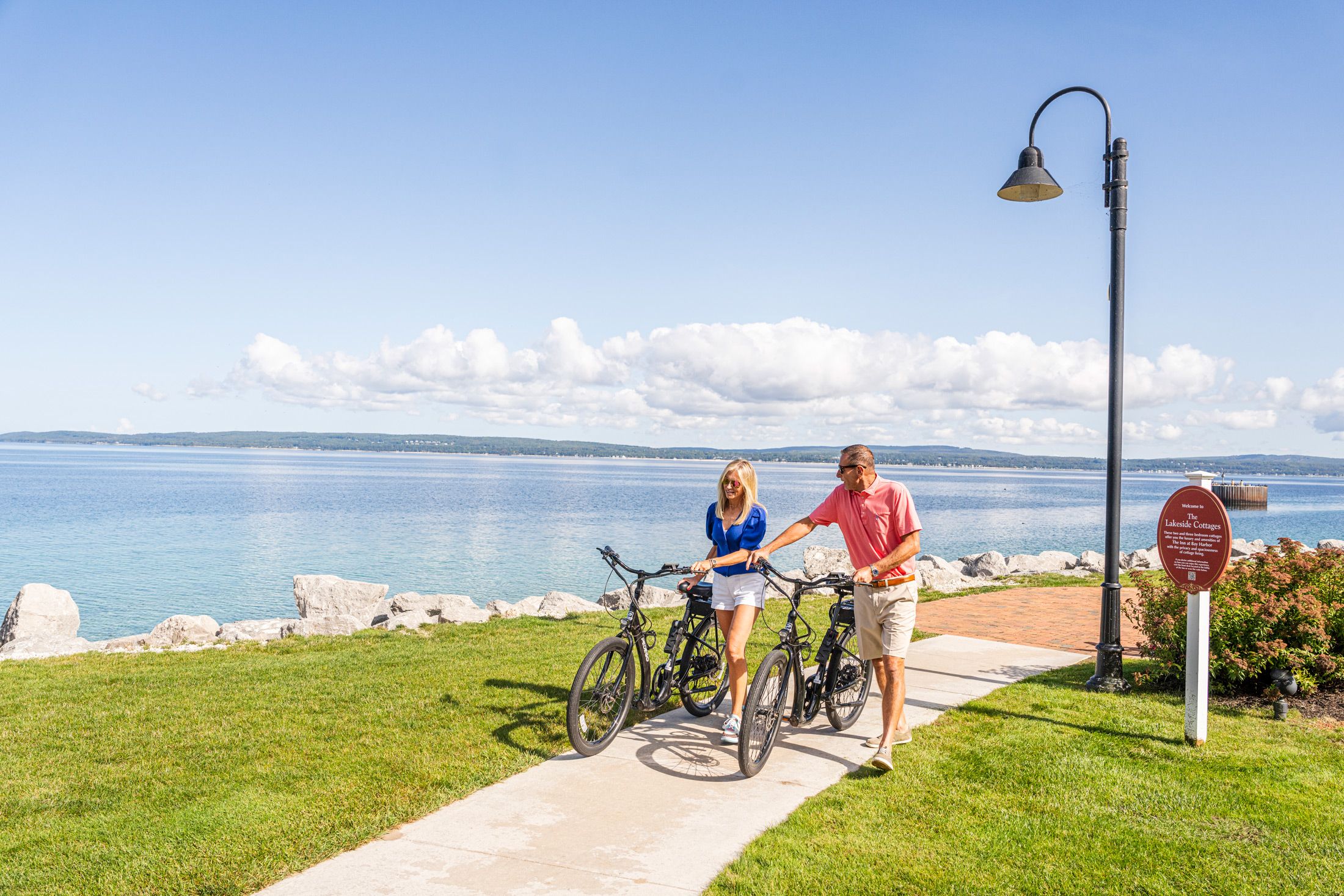 Couple in sunglasses walk bikes along waterfront path on sunny summer day