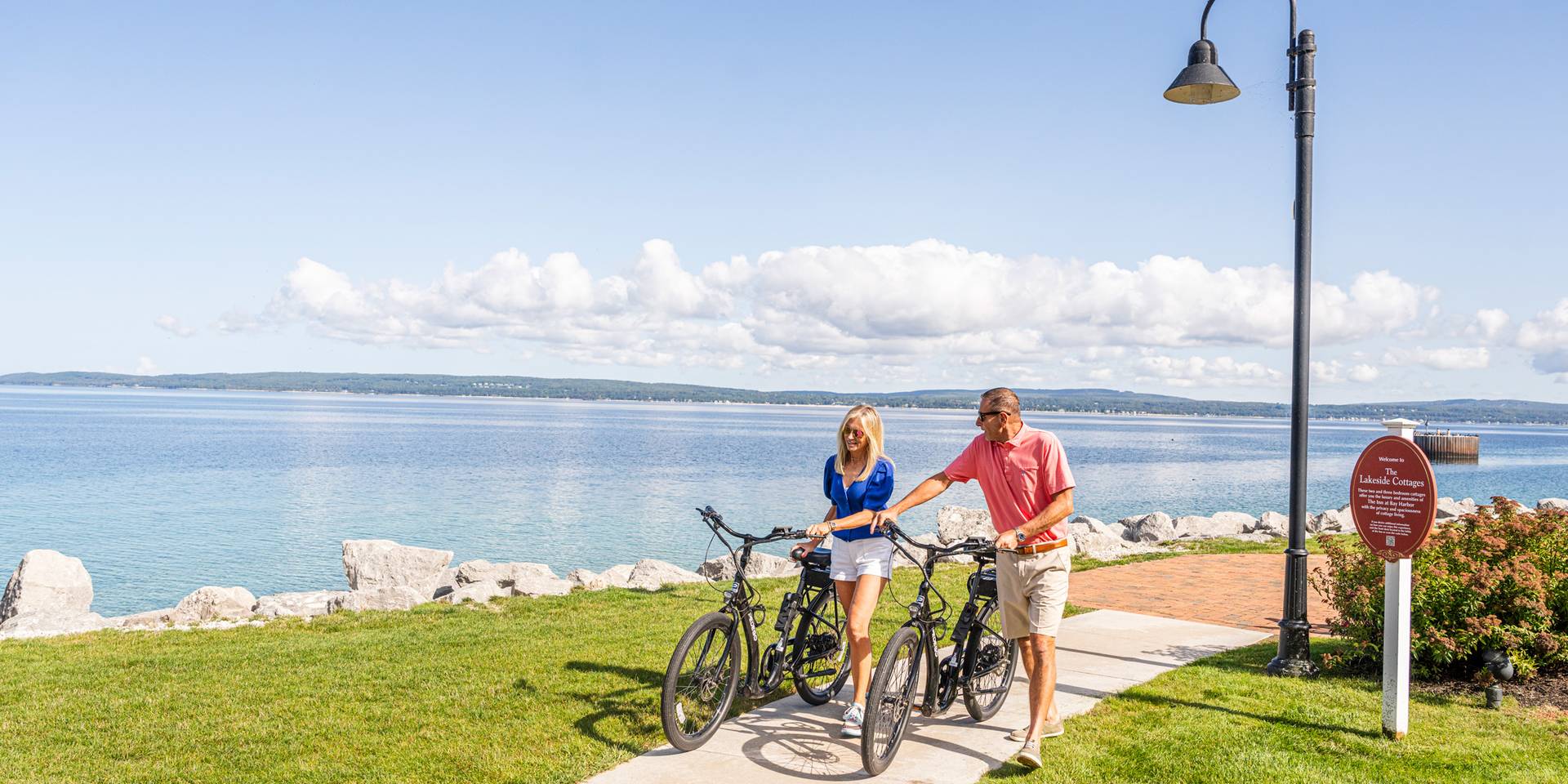 Couple in sunglasses walk bikes along waterfront path on sunny summer day