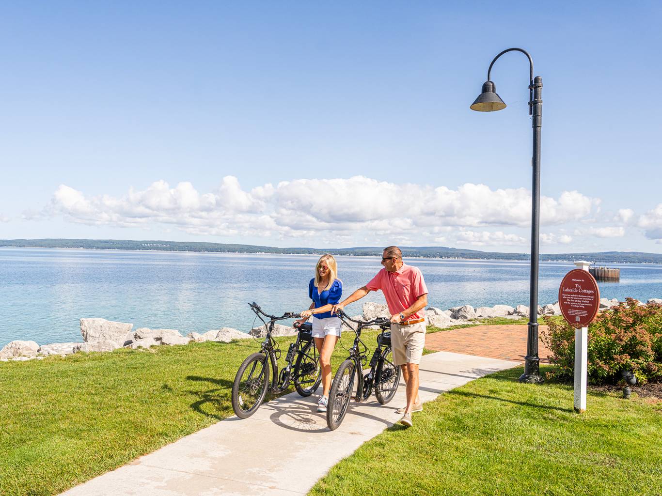 Couple in sunglasses walk bikes along waterfront path on sunny summer day