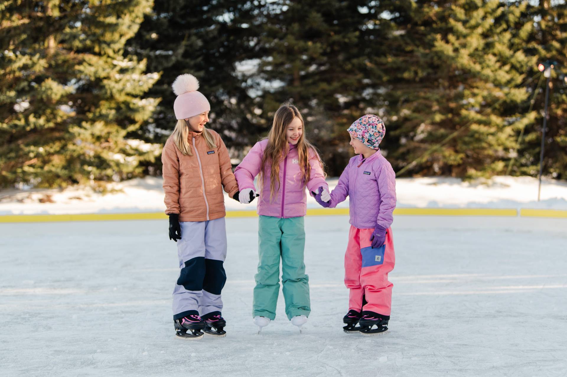 Smiling group of three young girls in bright winter gear, holding hands on outdoor ice rink