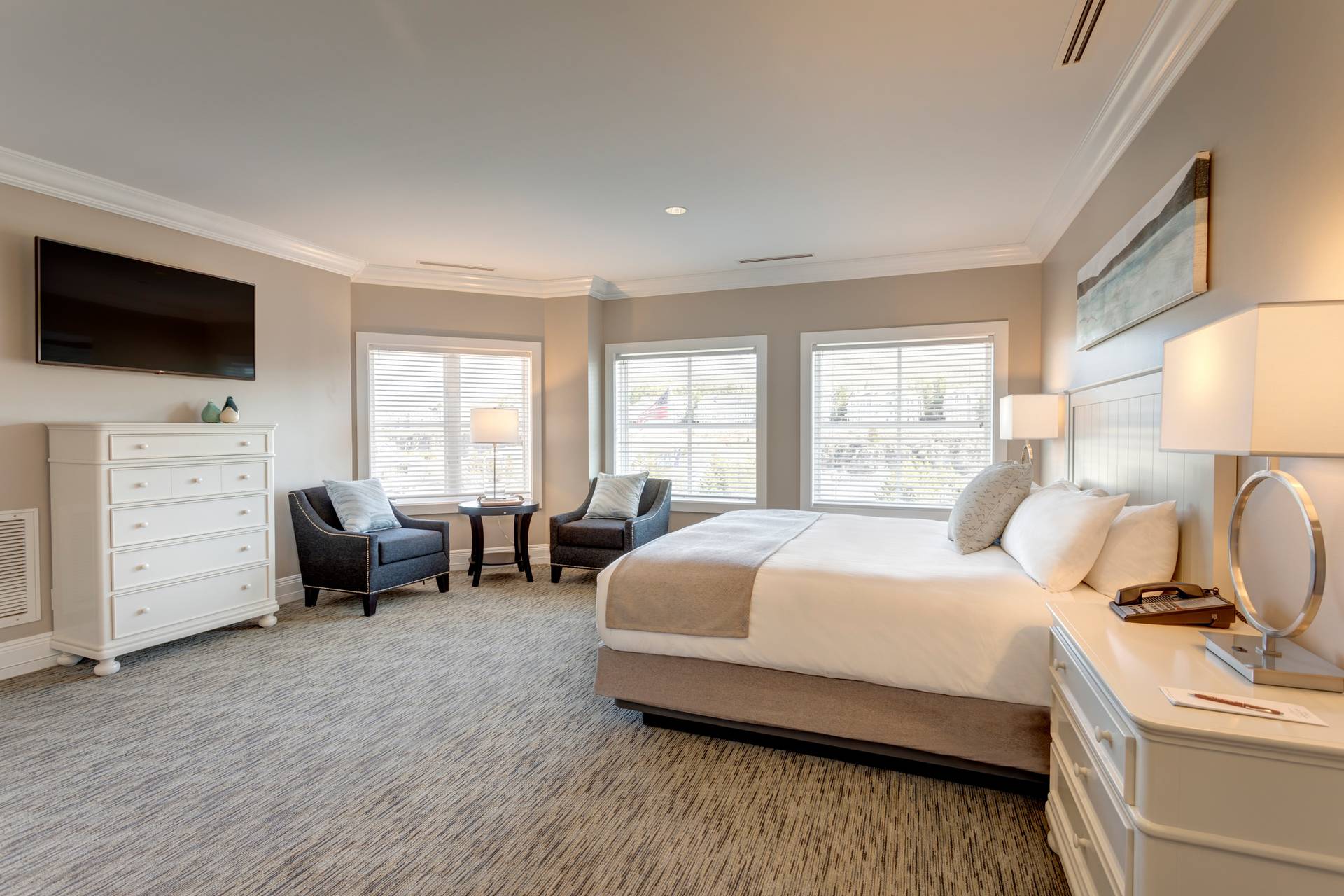 Wide view of Deluxe King hotel room with white bed, dresser, and chairs near large windows