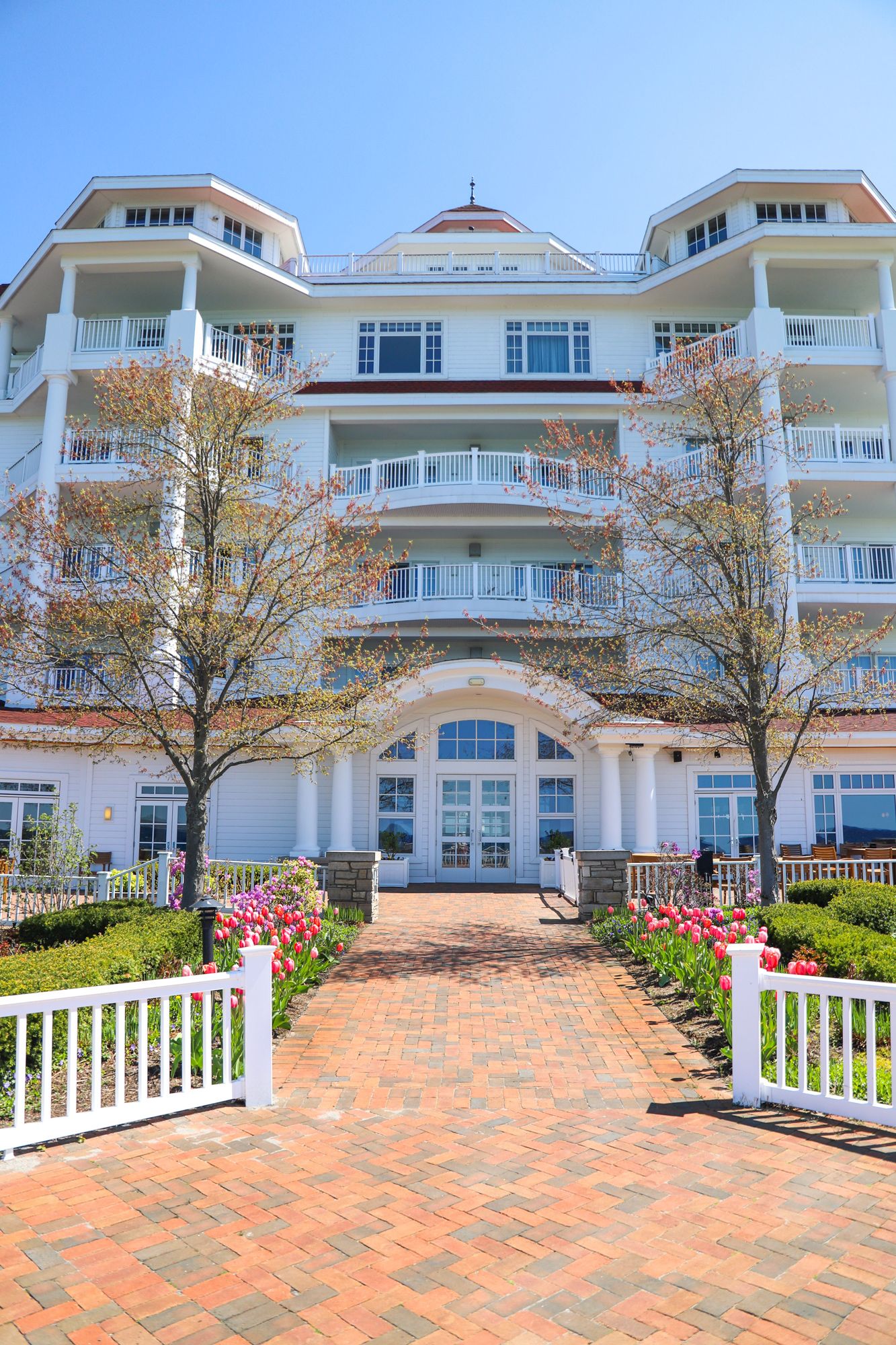 Arched entry of Inn at Bay Harbor with spring tulip gardens and budding trees, brick pathway, and classic lakefront architecture under a clear blue sky
