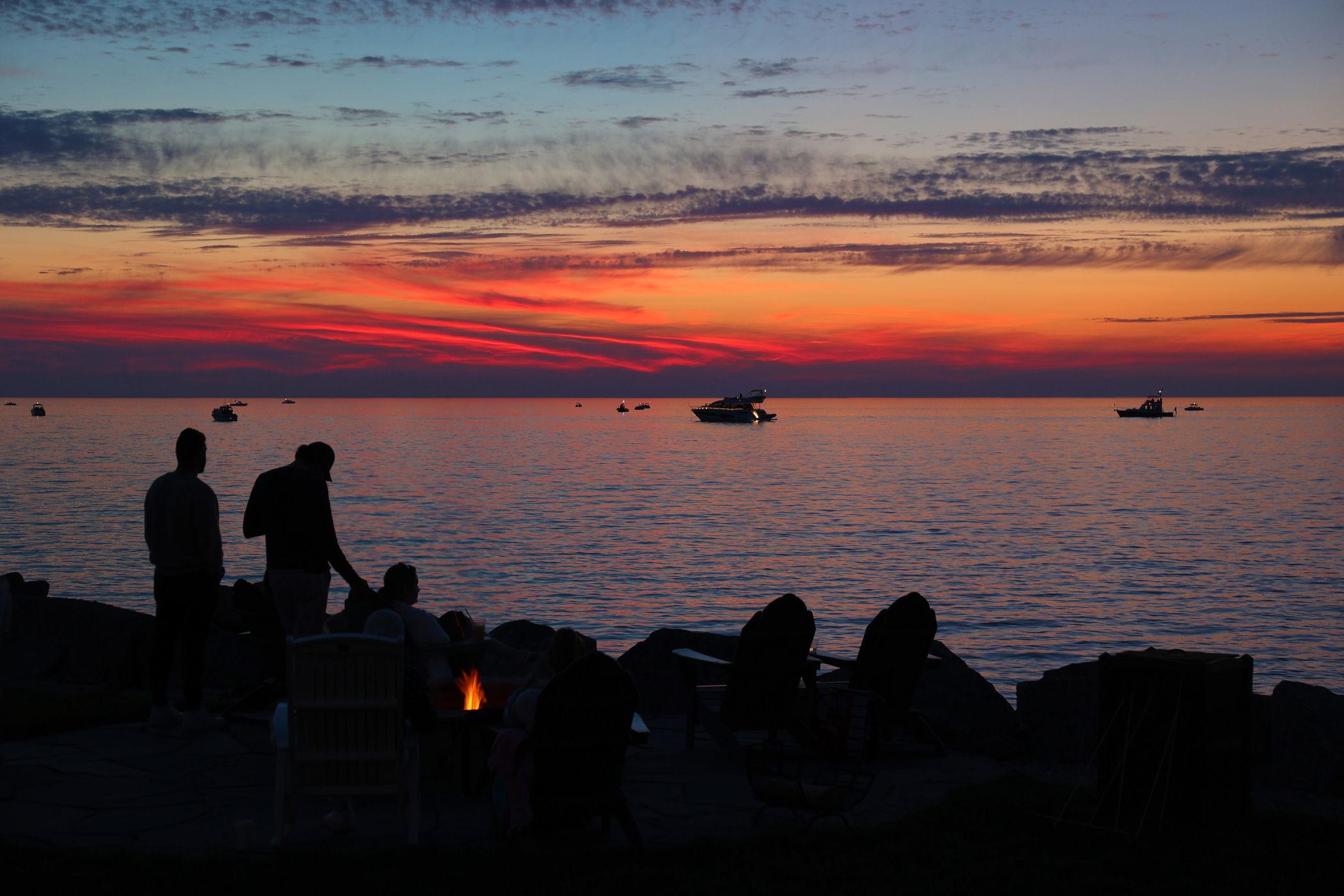 Shadows of people gathered by lakefront bonfire under red sunset watching boats on water