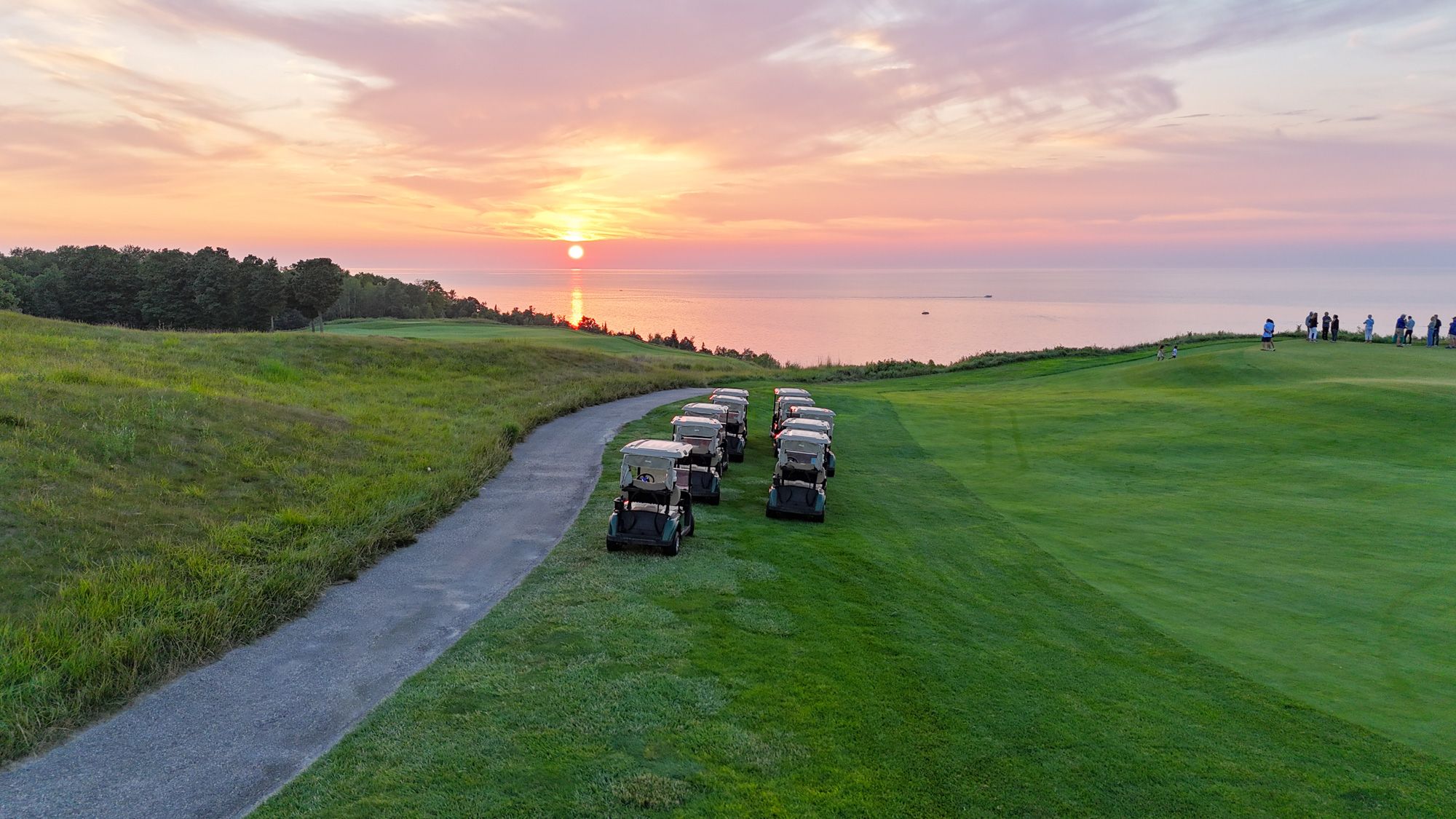 Golf carts line up next to path of golf course under orange and pink lakefront sunset