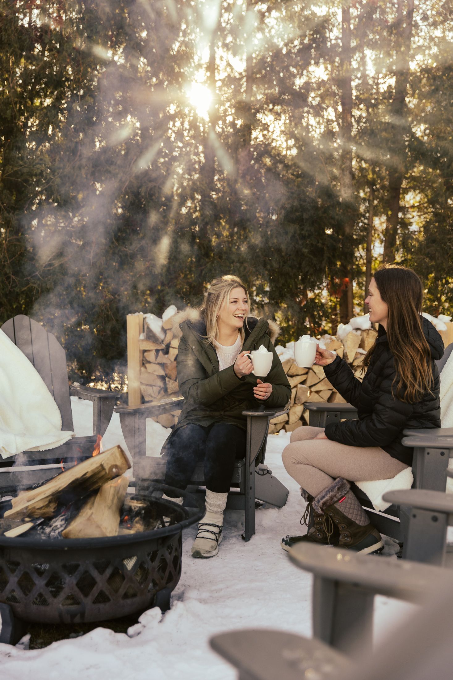 Two women chat by winter bonfire holding hot cocoa under filtered sunlight