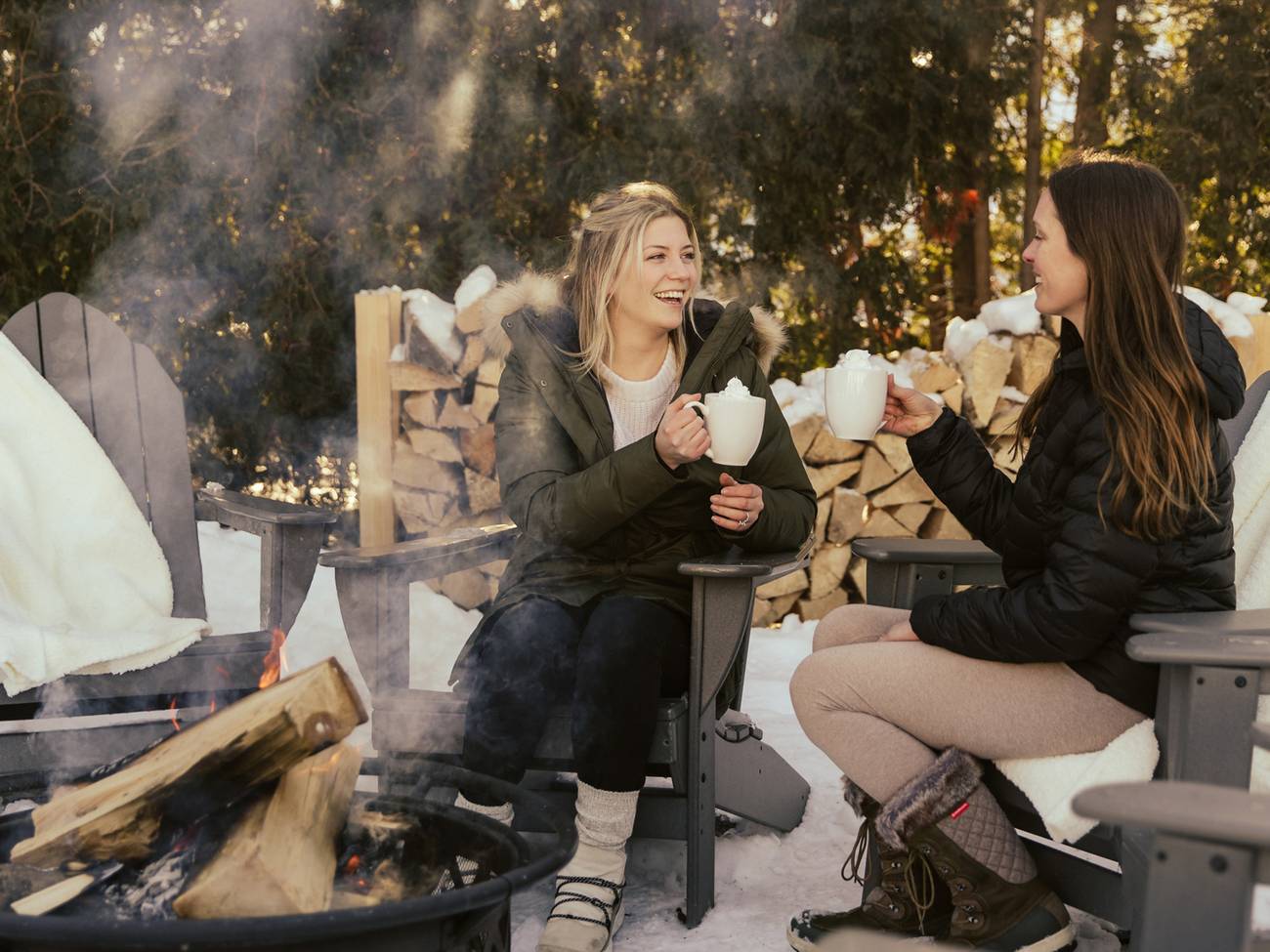 Two women chat by winter bonfire holding hot cocoa under filtered sunlight