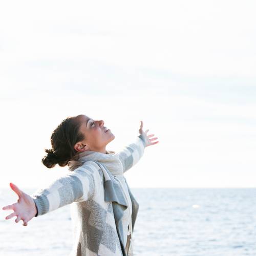 Woman smiles with open arms looking at sky in front of blue water