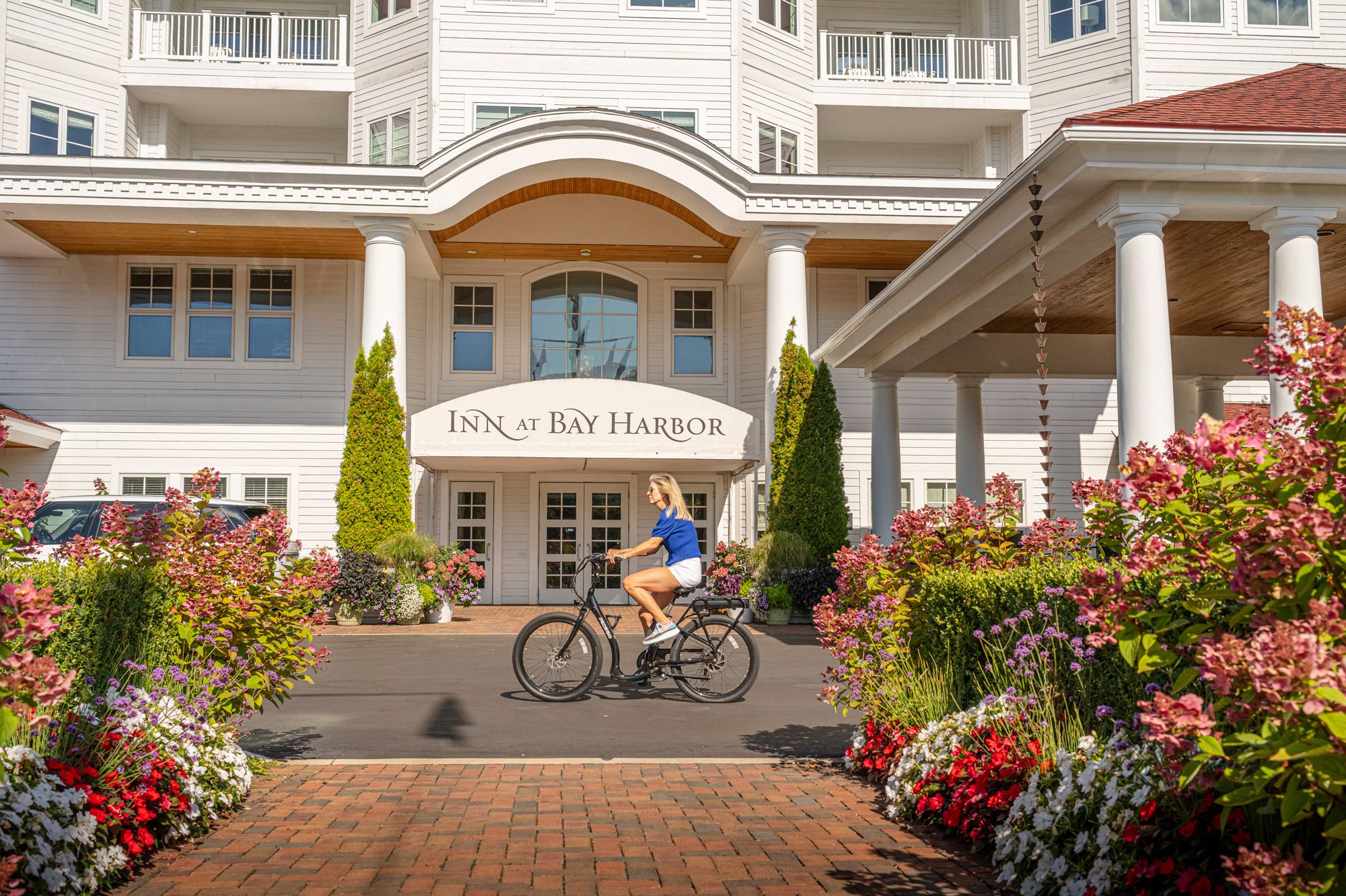 Blonde woman rides bicycle by colorful gardens at front entrance of Inn at Bay Harbor