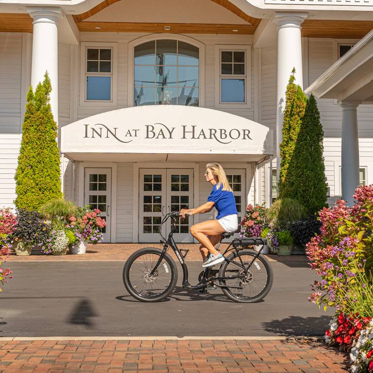 Blonde woman rides bicycle by colorful gardens at front entrance of Inn at Bay Harbor