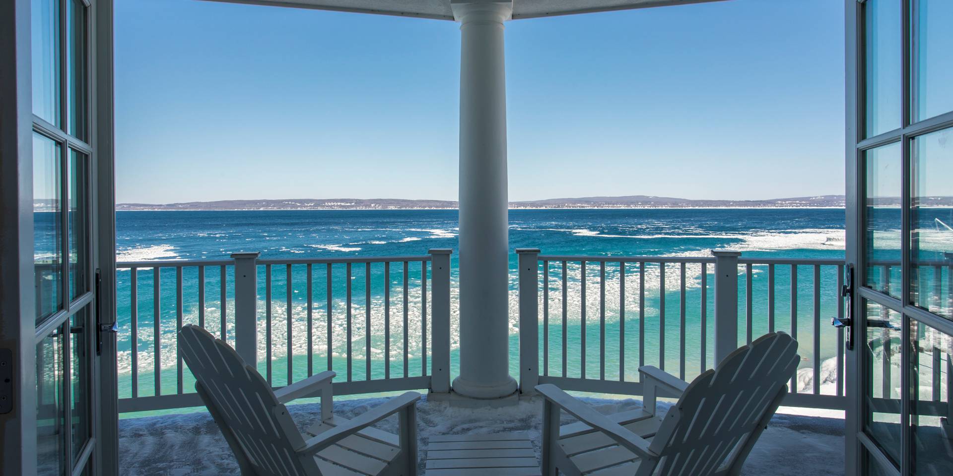 Wide winter balcony with open French doors, white Adirondack chairs, overlooking bright, icy Lake Michigan under blue skies
