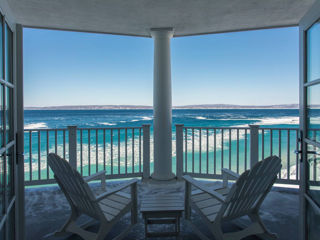 Wide winter balcony with open French doors, white Adirondack chairs, overlooking bright, icy Lake Michigan under blue skies