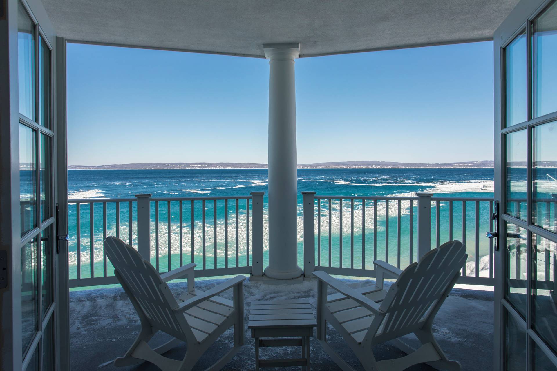 Wide winter balcony with open French doors, white Adirondack chairs, overlooking bright, icy Lake Michigan under blue skies