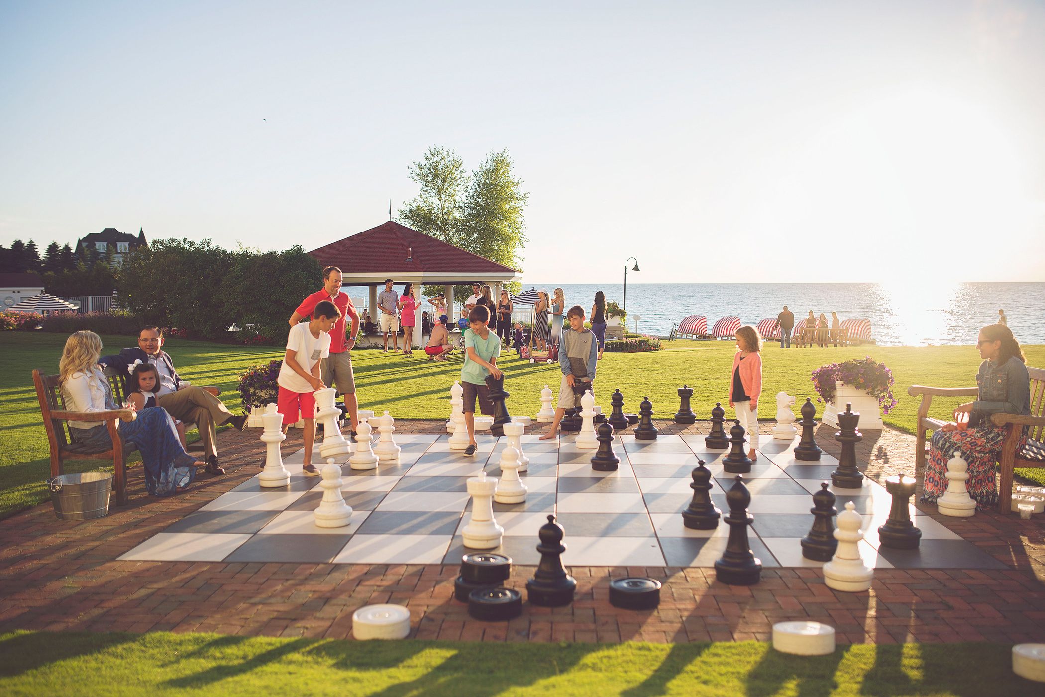 Two families play giant chess outdoors on bustling lakefront lawn as sun sets over water