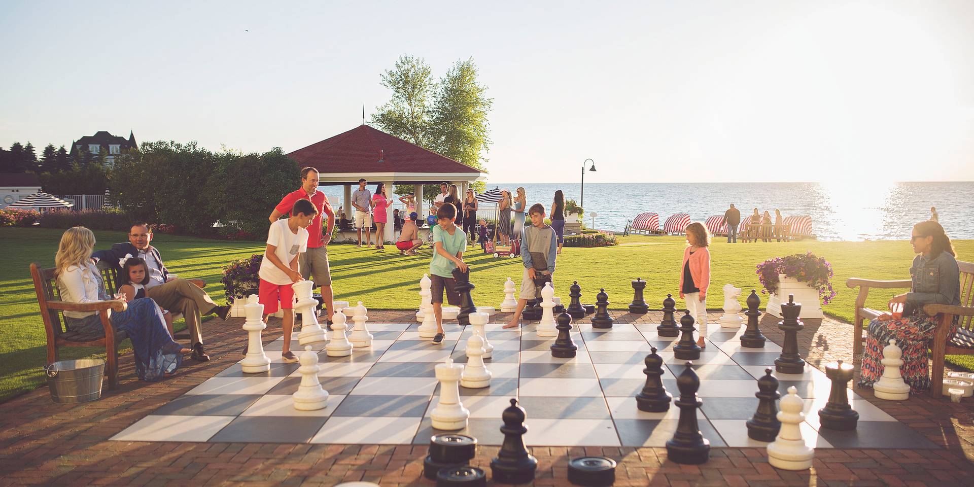 Two families play giant chess outdoors on bustling lakefront lawn as sun sets over water