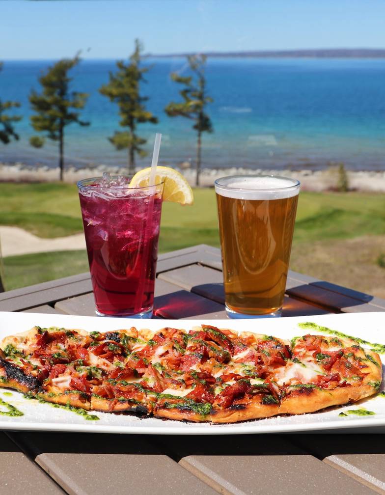 Two drinks and colorful flatbread on outdoor table overlooking Quarry course and water, Bay Harbor Golf Club