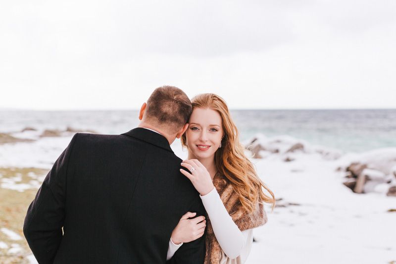 Red haired bride smiles as she embraces groom near snowy lakefront