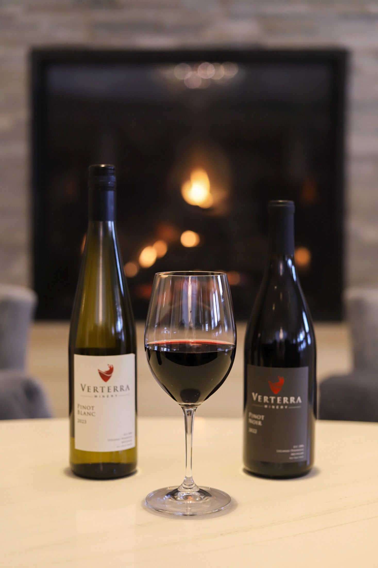 Glass of red wine, between Verterra red, white wine bottles near fireplace