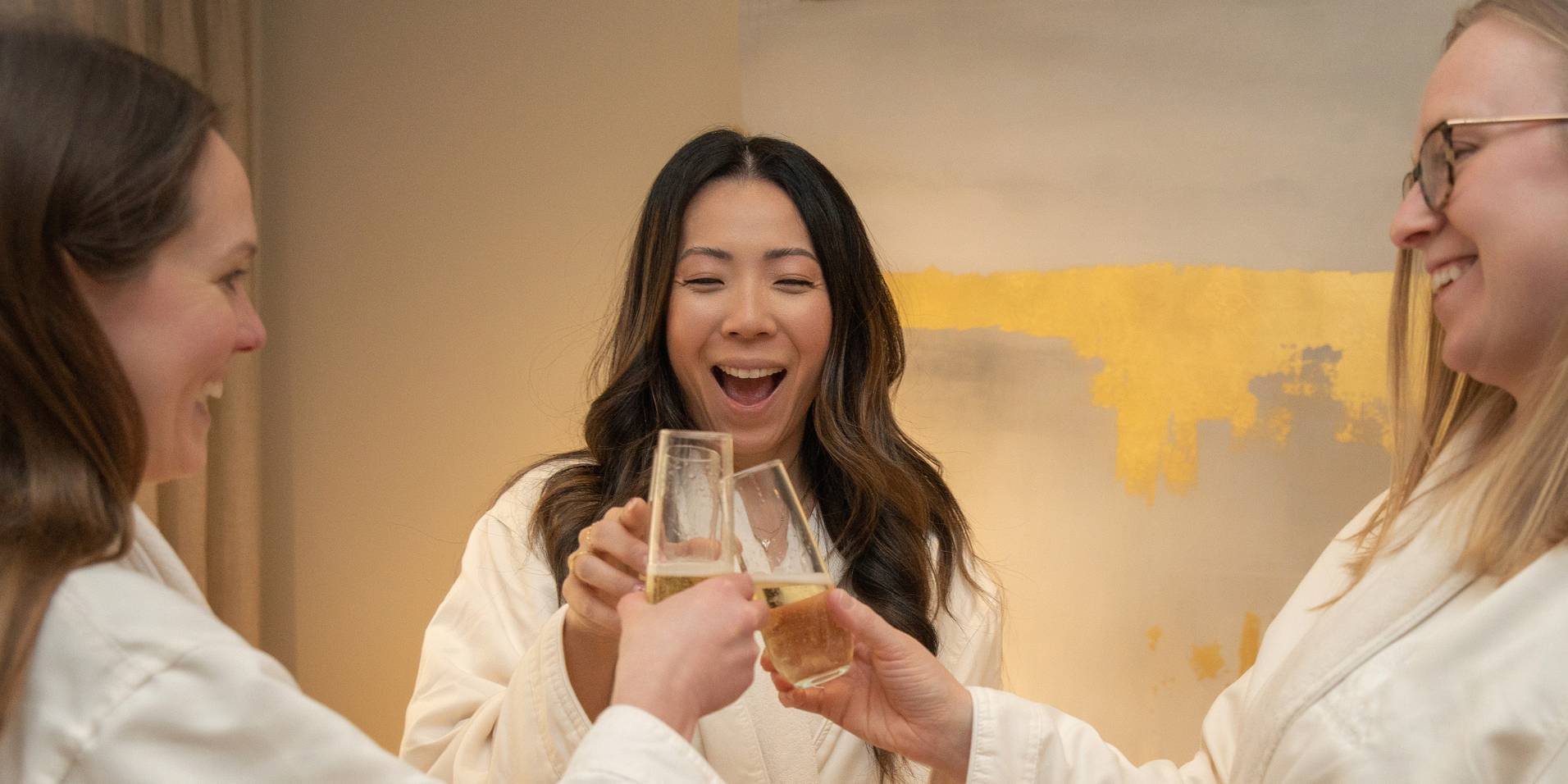 Three laughing women friends in white spa robes toast champagne flutes, The Spa