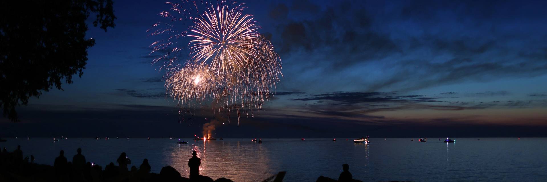 Gold fireworks light up the water as shadows of people watch under darkening sky, Bay Harbor