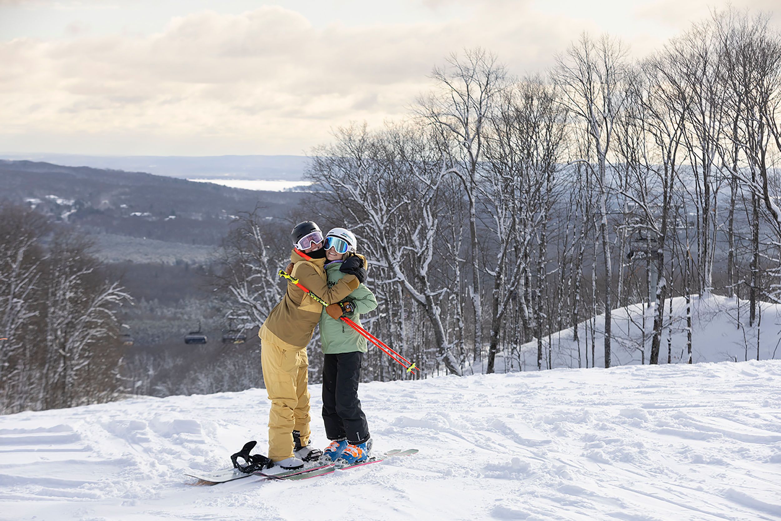 Two girls in ski/snowboard gear hugging on top of the slopes