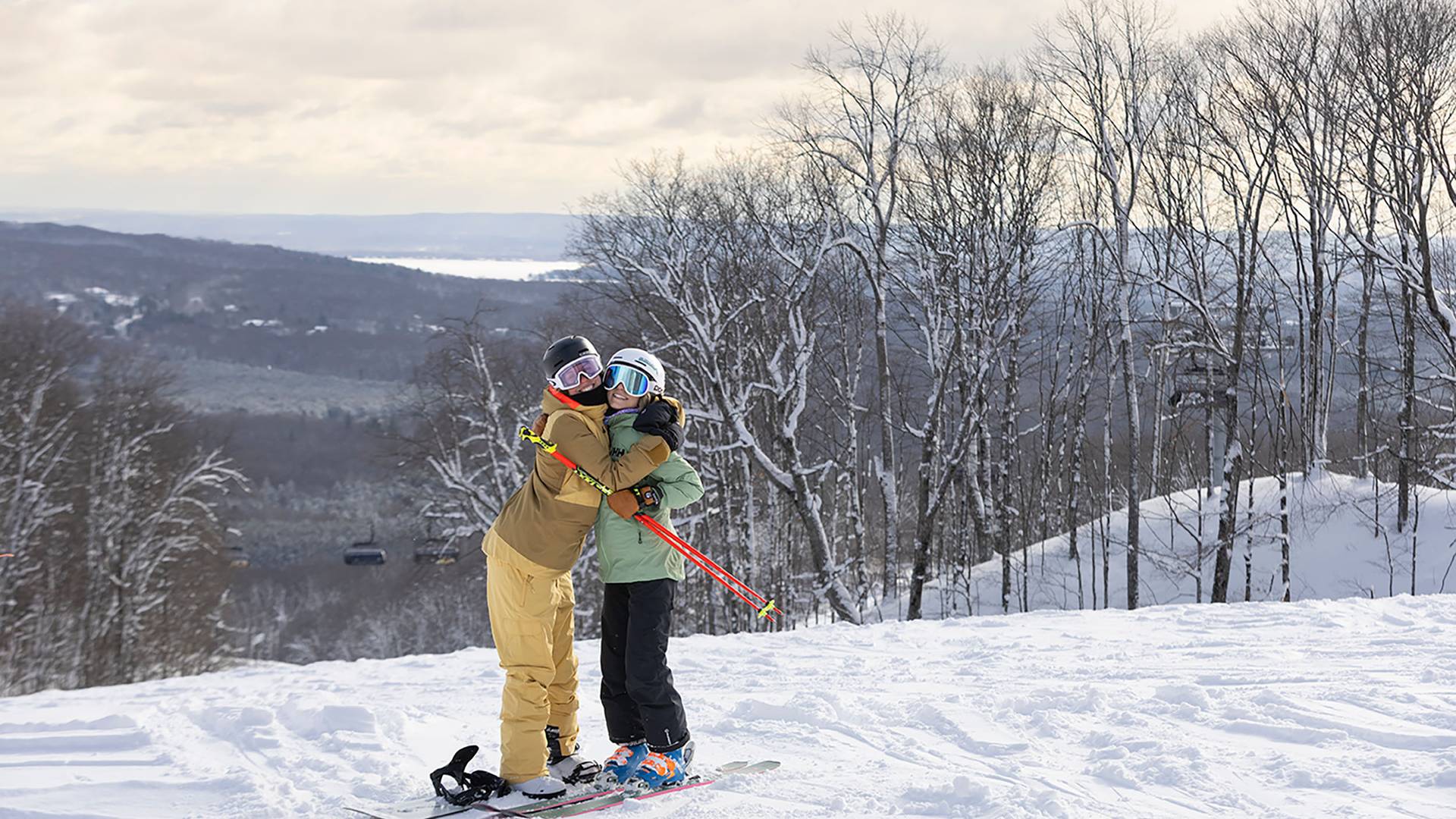Two girls in ski/snowboard gear hugging on top of the slopes