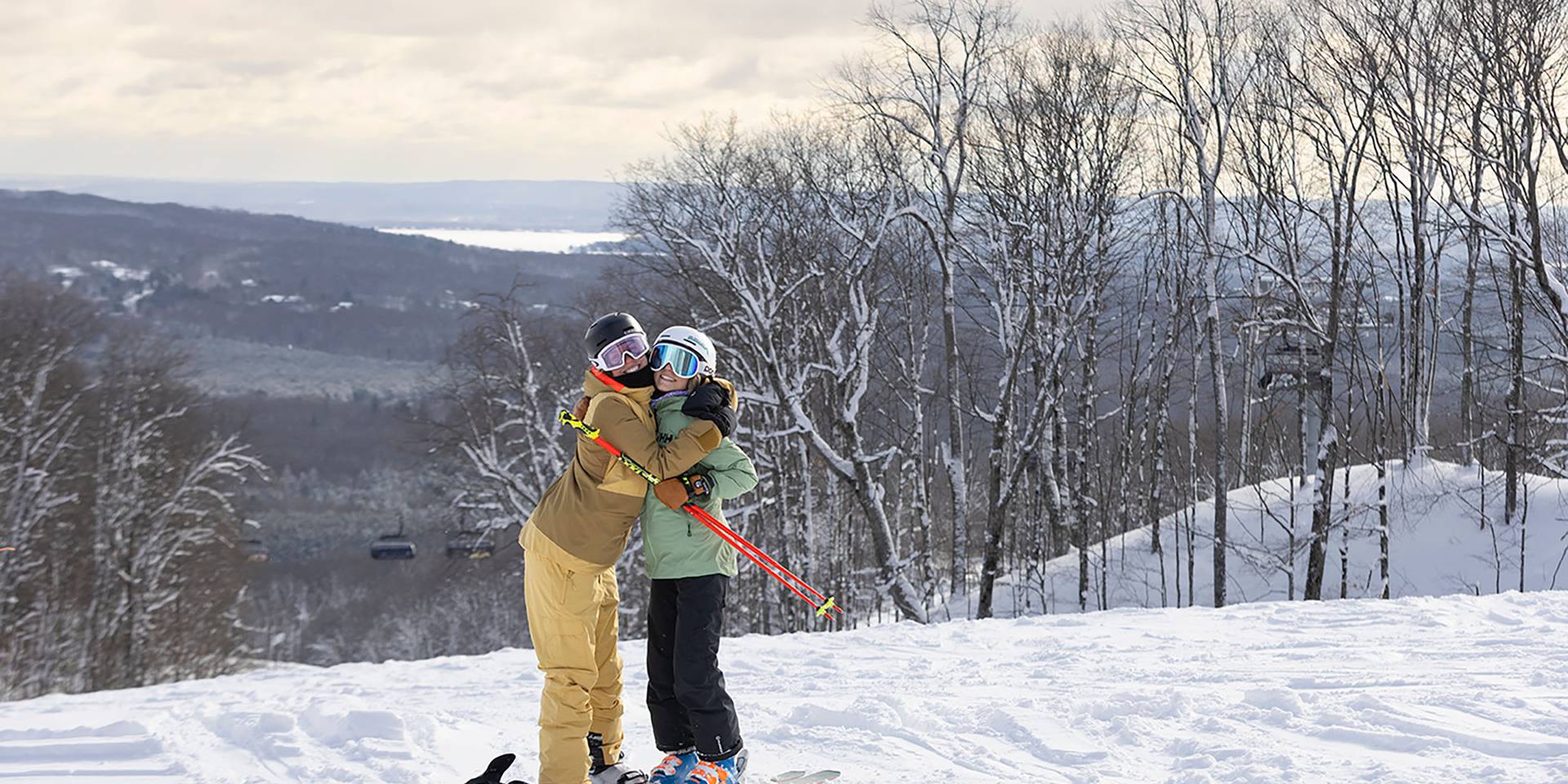 Two girls in ski/snowboard gear hugging on top of the slopes