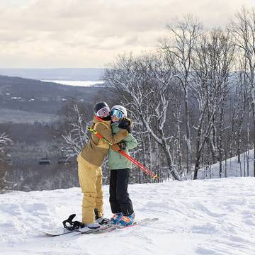 Two girls in ski/snowboard gear hugging on top of the slopes