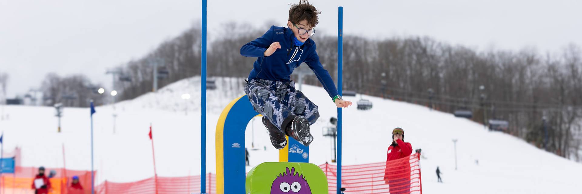 A kid in ski boats and winter gear jumping over a sign
