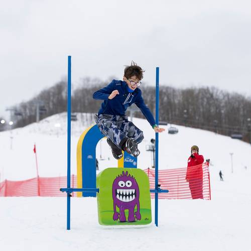 A kid in ski boats and winter gear jumping over a sign
