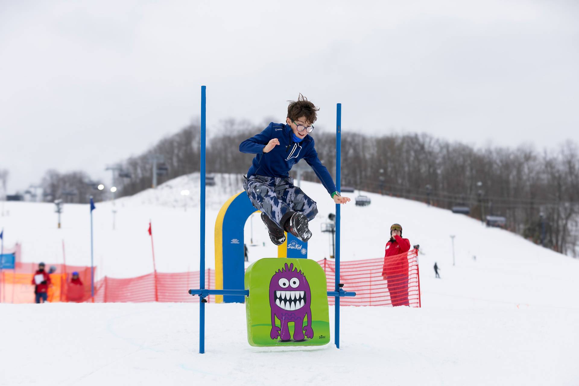 A kid in ski boats and winter gear jumping over a sign