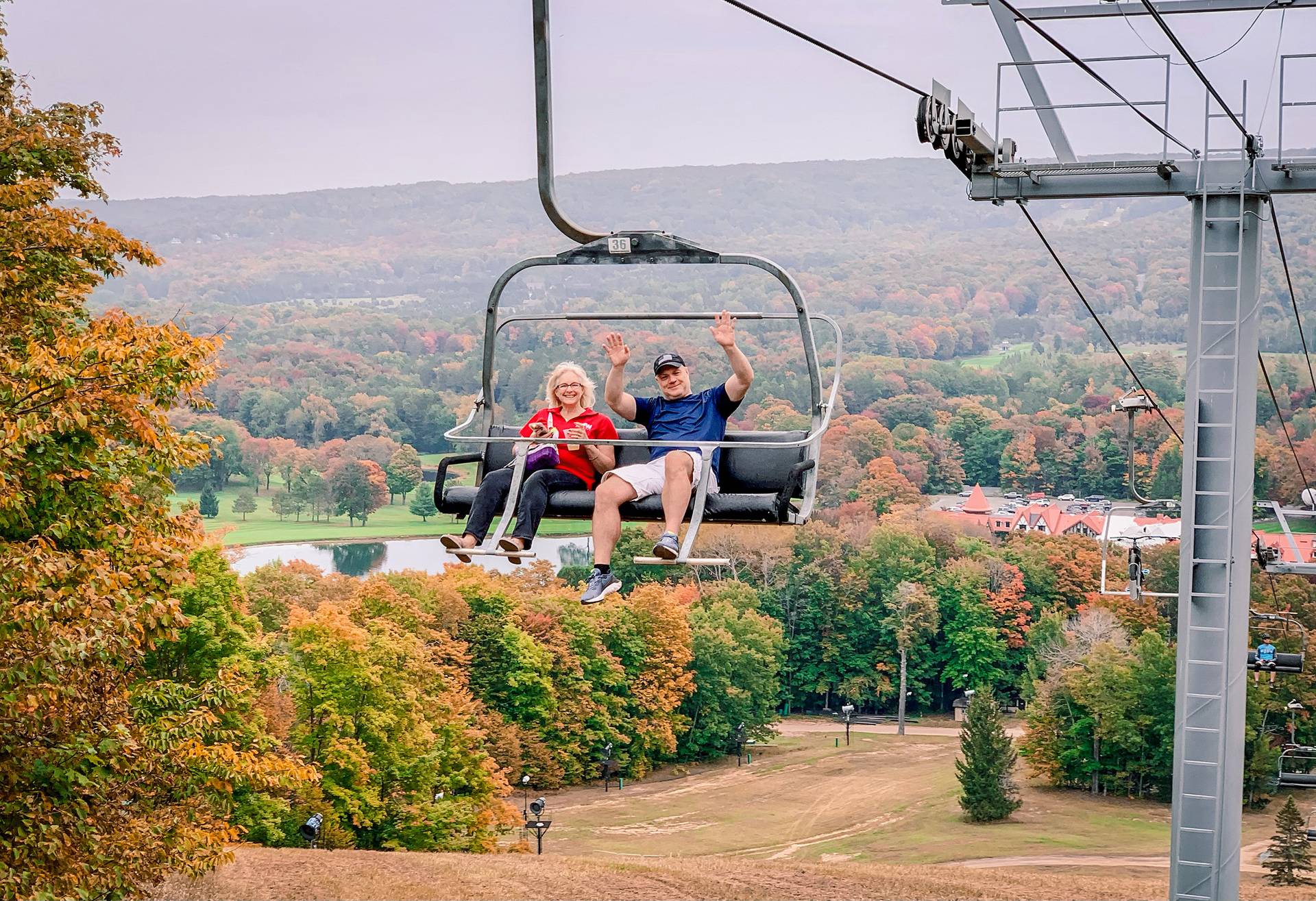 A couple smiling and waving on a chairlift during fall