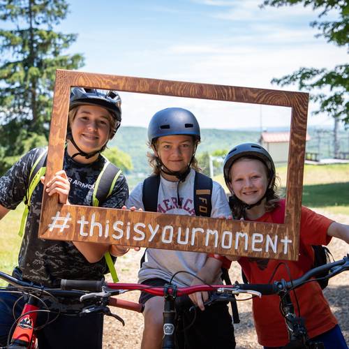 A group of kids holding a wooden frame sign smiling on mountain bikes