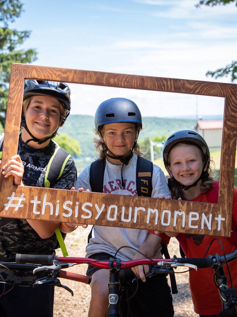 A group of kids holding a wooden frame sign smiling on mountain bikes