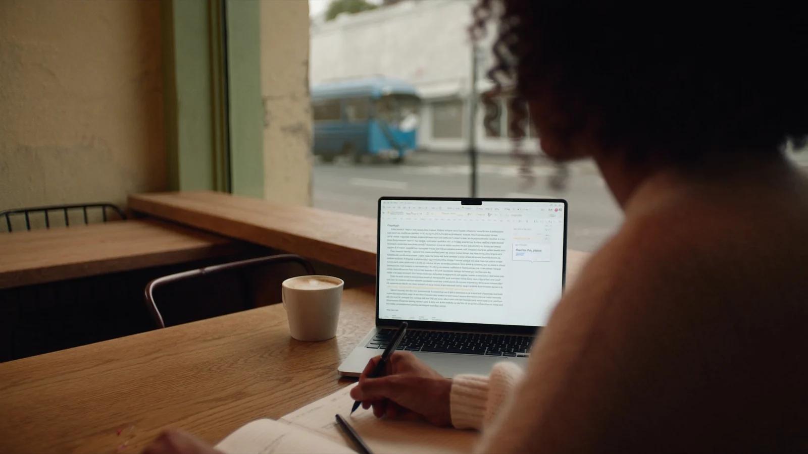 Frau schreibt am Laptop im Café einen Absatz neu, daneben liegt ein Notizbuch