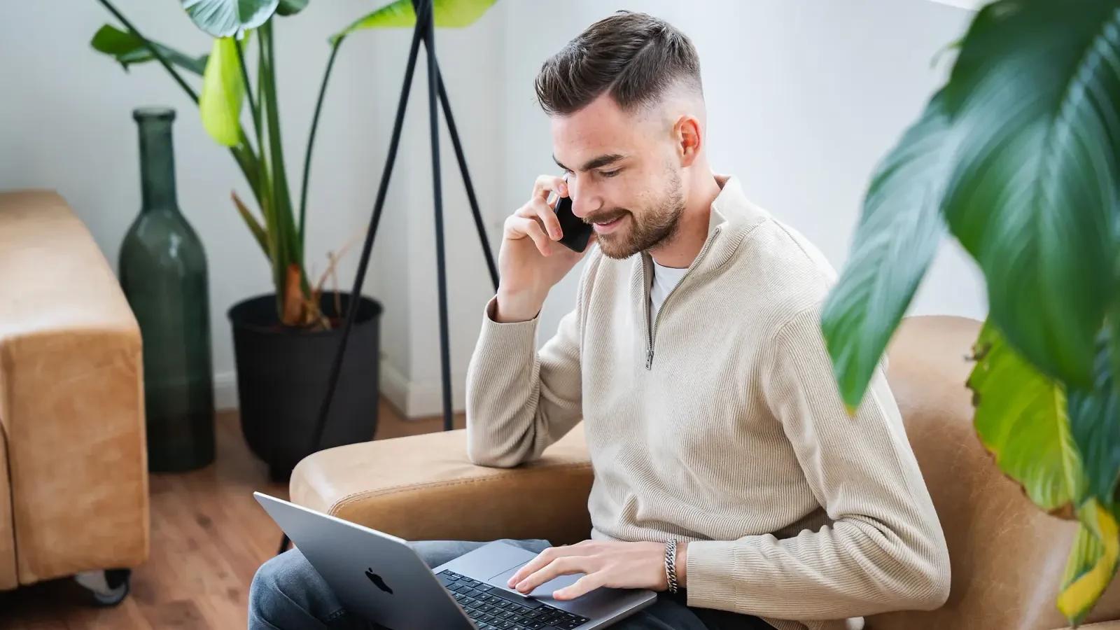 Jan Philipps im hellen Pullover telefoniert auf einem Sessel, den Laptop auf dem Schoß