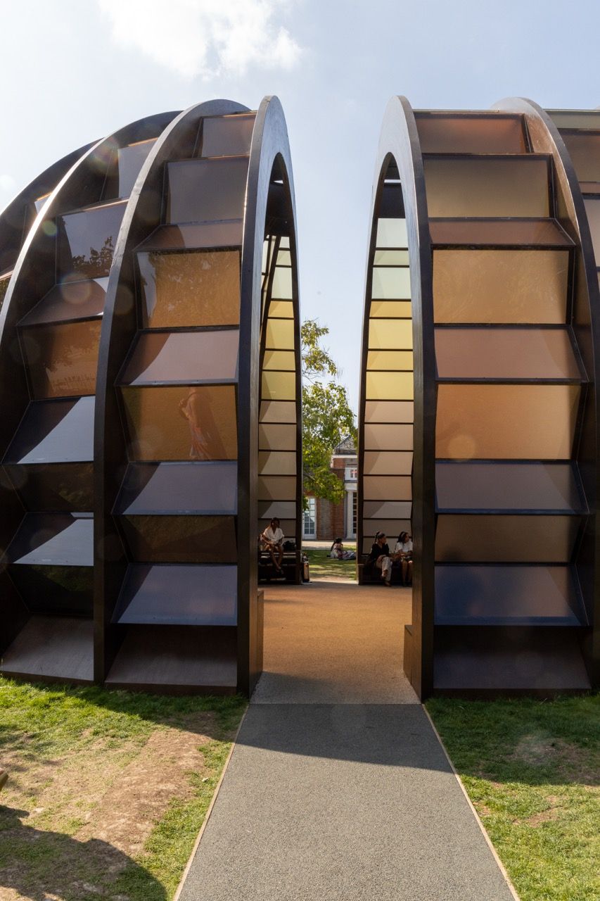 A wide-angle view of the Serpentine Pavilion in a park setting, showing its modern, curved design against a backdrop of trees and a blue sky.