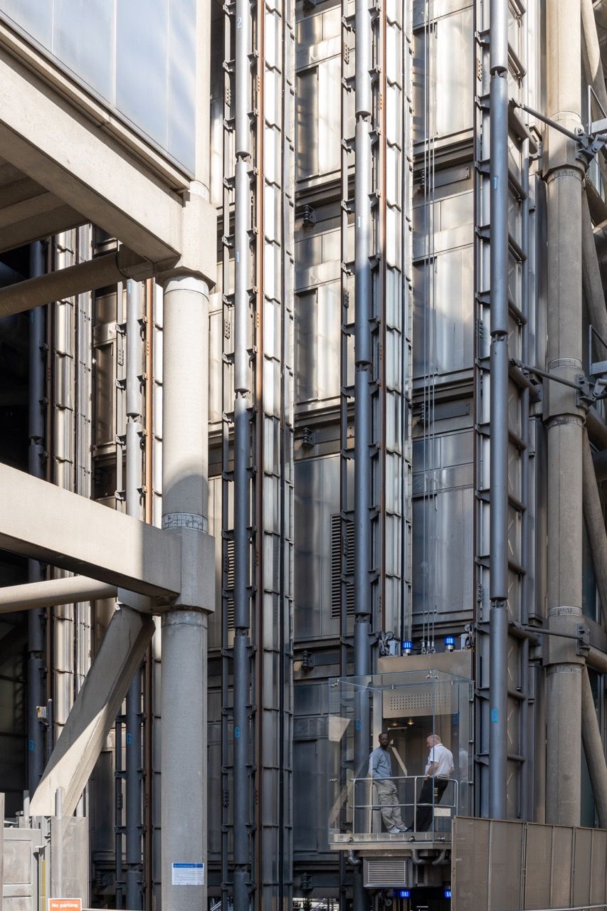 A wide-angle, exterior view of the **Lloyd's Building** in London, showcasing its distinctive inside-out architecture with exposed pipes, ducts, and service elements on the facade. The building stands tall against the skyline, reflecting the surrounding urban environment.