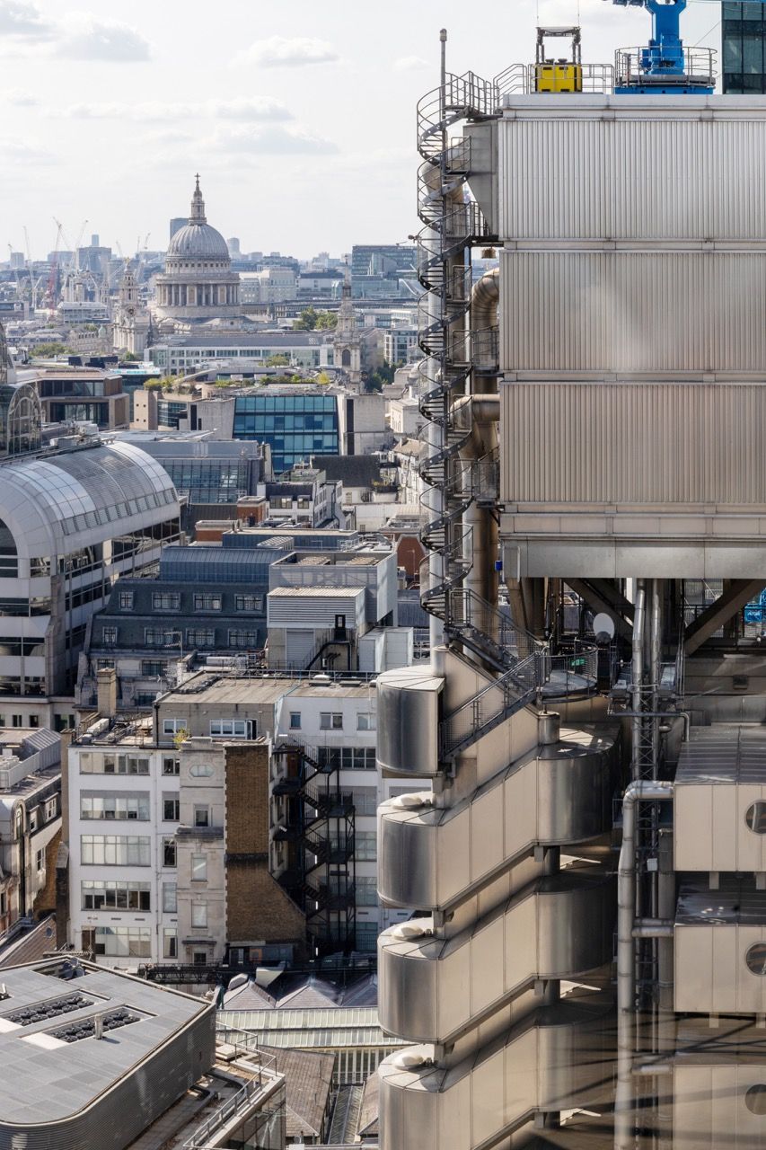 A wide-angle, exterior view of the **Lloyd's Building** in London, showcasing its distinctive inside-out architecture with exposed pipes, ducts, and service elements on the facade. The building stands tall against the skyline, reflecting the surrounding urban environment.