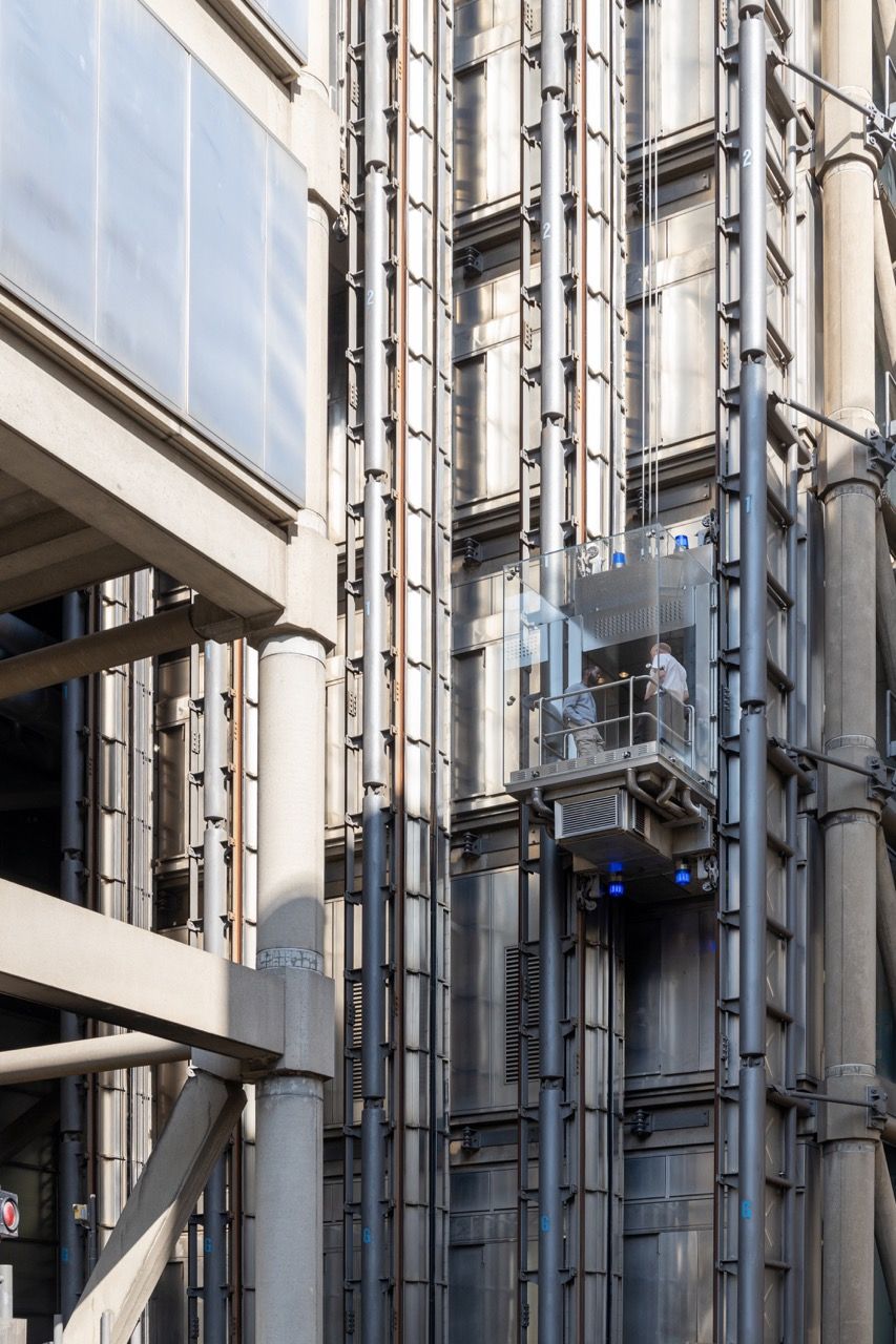 A wide-angle, exterior view of the **Lloyd's Building** in London, showcasing its distinctive inside-out architecture with exposed pipes, ducts, and service elements on the facade. The building stands tall against the skyline, reflecting the surrounding urban environment.