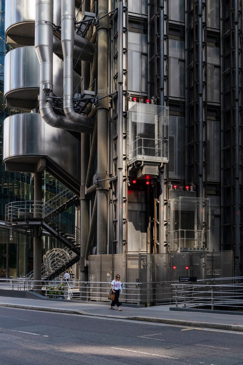 A wide-angle, exterior view of the **Lloyd's Building** in London, showcasing its distinctive inside-out architecture with exposed pipes, ducts, and service elements on the facade. The building stands tall against the skyline, reflecting the surrounding urban environment.