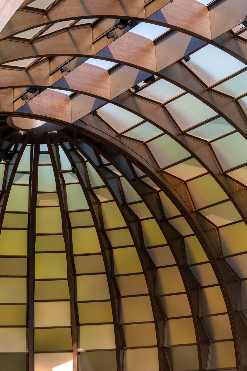 A wide-angle view of the Serpentine Pavilion in a park setting, showing its modern, curved design against a backdrop of trees and a blue sky.
