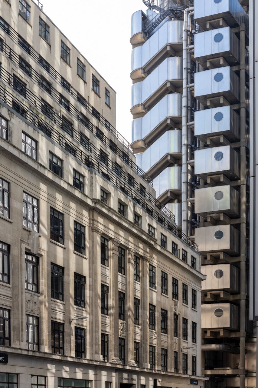 A wide-angle, exterior view of the **Lloyd's Building** in London, showcasing its distinctive inside-out architecture with exposed pipes, ducts, and service elements on the facade. The building stands tall against the skyline, reflecting the surrounding urban environment.