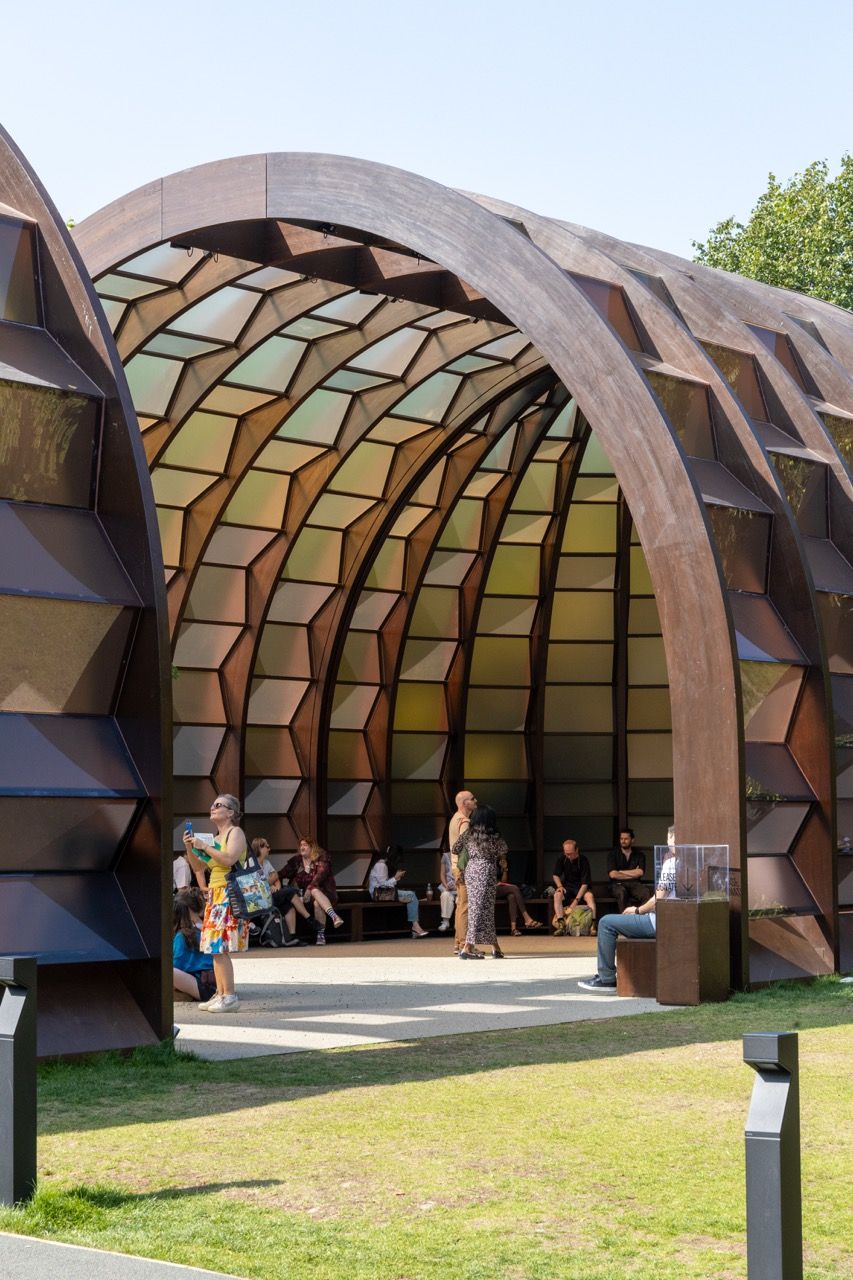 A wide-angle view of the Serpentine Pavilion in a park setting, showing its modern, curved design against a backdrop of trees and a blue sky.