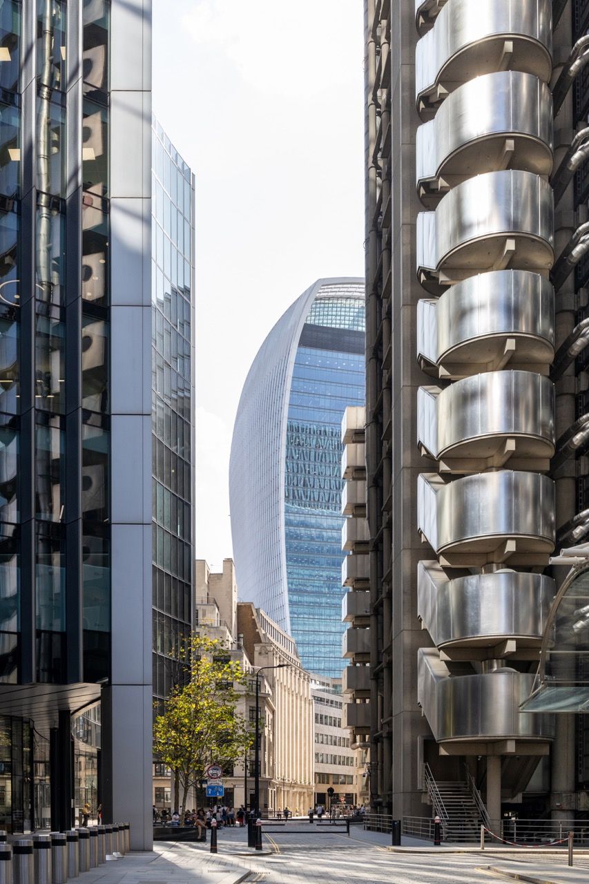 A wide-angle, exterior view of the **Lloyd's Building** in London, showcasing its distinctive inside-out architecture with exposed pipes, ducts, and service elements on the facade. The building stands tall against the skyline, reflecting the surrounding urban environment.