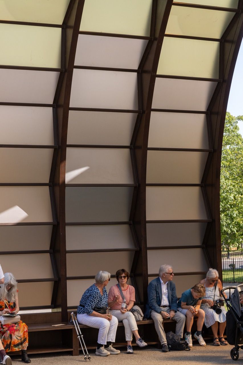 A wide-angle view of the Serpentine Pavilion in a park setting, showing its modern, curved design against a backdrop of trees and a blue sky.