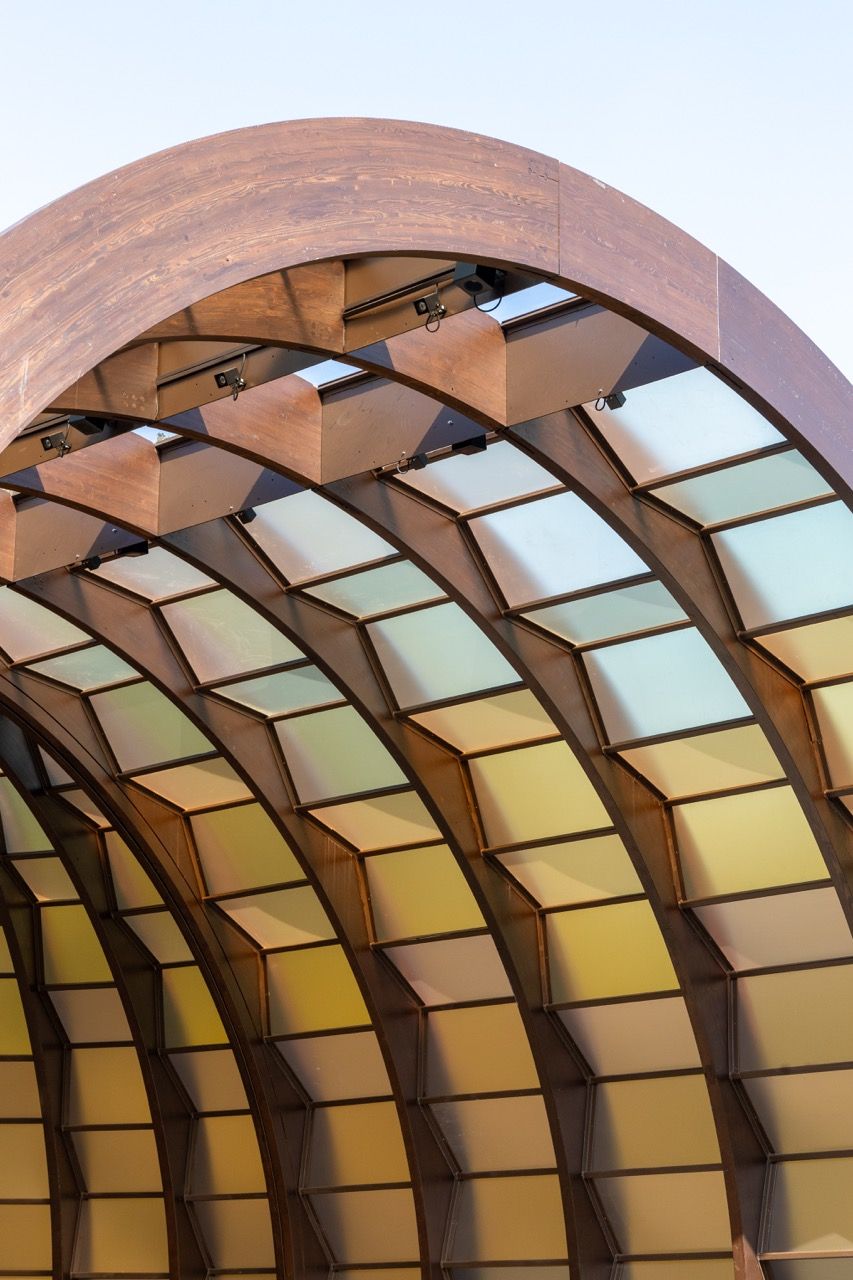 A wide-angle view of the Serpentine Pavilion in a park setting, showing its modern, curved design against a backdrop of trees and a blue sky.