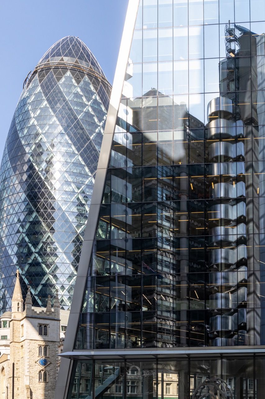 A wide-angle, exterior view of the **Lloyd's Building** in London, showcasing its distinctive inside-out architecture with exposed pipes, ducts, and service elements on the facade. The building stands tall against the skyline, reflecting the surrounding urban environment.