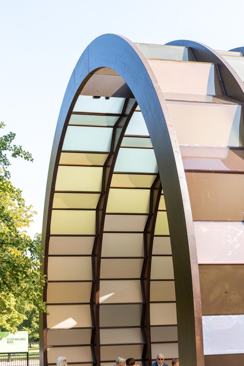 A wide-angle view of the Serpentine Pavilion in a park setting, showing its modern, curved design against a backdrop of trees and a blue sky.